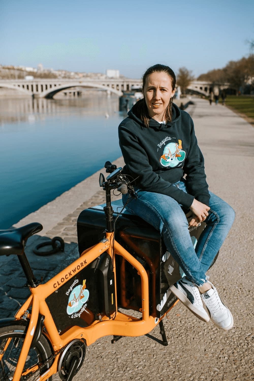 Femme en sweat Baguette à Bicyclette assise sur un vélo cargo orange de la marque, sur les quais à Lyon